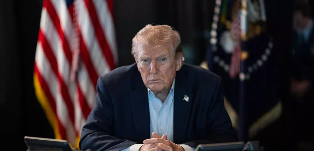 Donald Trump, presidente dos Estados Unidos, sentado à mesa diante de bandeiras americanas.