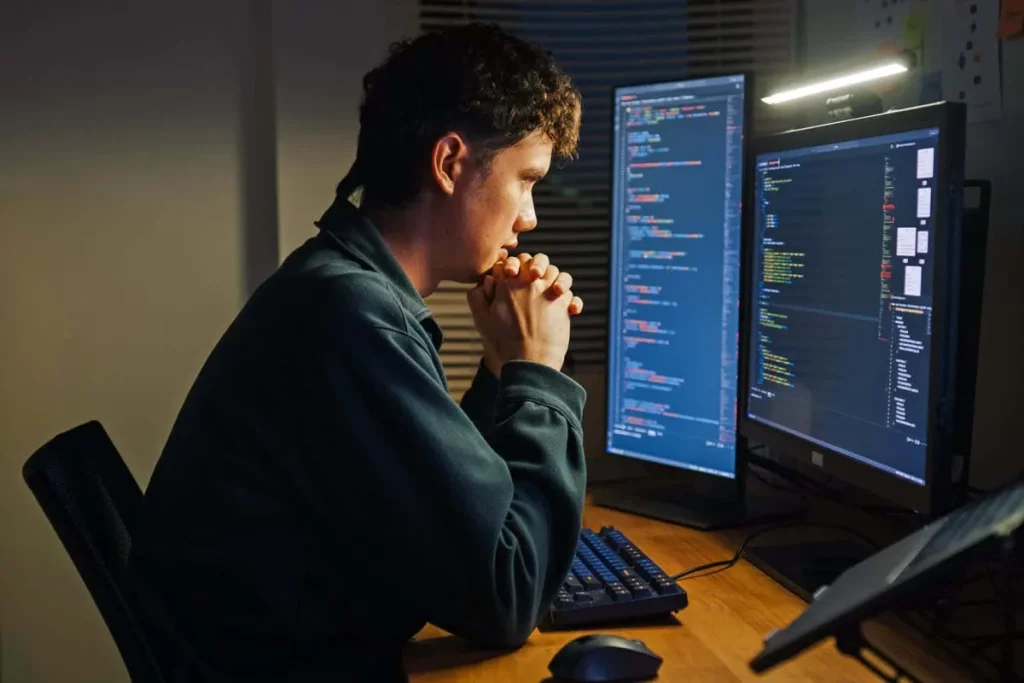 Desenvolvedor trabalha em código em frente a monitores com programação exibida. (Foto: newscientist.com)
