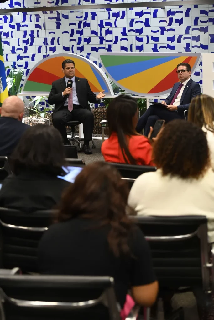 Um homem em traje formal discursa durante evento com painel colorido ao fundo, enquanto público o acompanha. (Foto: Wikimedia